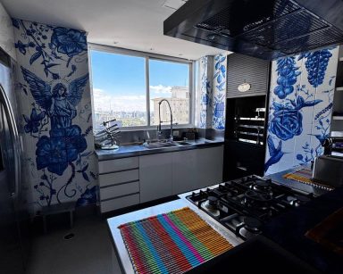 nspirada na tradição do azulejo português, a parede desta cozinha ganhou uma composição em azul cobalto sobre fundo branco que mescla frutos, santos, beija-flores e vegetação. A técnica resgata a estética clássica dos revestimentos ibéricos, mas a escolha dos elementos e o desenho trazem uma identidade própria — criando um frescor decorativo que dialoga com o cotidiano do lar sem perder a sofisticação artesanal da pintura sobre azulejo.